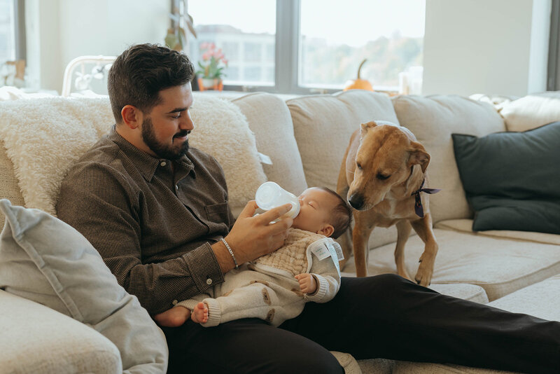 father feeding baby with dog looking on during in home newborn photos captured by NYC newborn photographer Elsie Goodman 