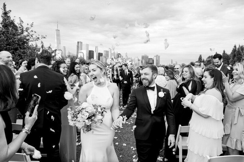 bride and groom excitedly holding hands walking down the aisle after getting married with guests throwing flower petals outside of liberty house in jersey city
