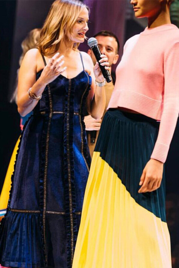 Creative director Katy Reeve presenting on stage at a hair awards event, standing beside a model in a pleated skirt and pink sweater.