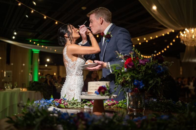 Bride and groom sharing cake during their wedding reception at the White Room in historic downtown St. Augustine. The bride in a white lace dress feeds cake to the groom in a navy suit, surrounded by string lights and colorful floral arrangements with red, purple, and blue flowers.