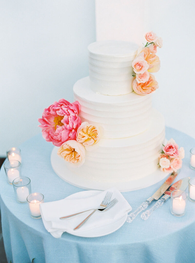 Newport Wedding Photographer | Three-tier white wedding cake adorned with pink and peach flowers on a blue table. Surrounded by candles, with a plate and cutlery beside it.