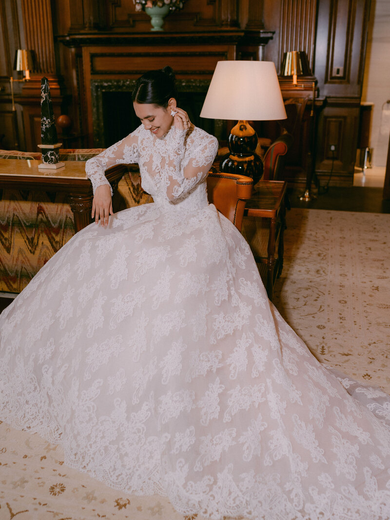 Bride in the Churchill Suite at Raffles at The OWO in London, wearing a Monique Lhuillier lace bridal gown, elegantly seated on a chair.