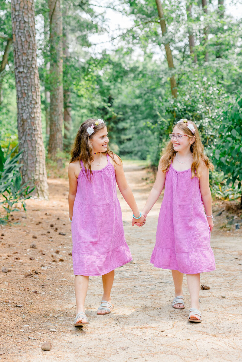 Sisters in pink dresses holding hands walking down a wooded path with greenery