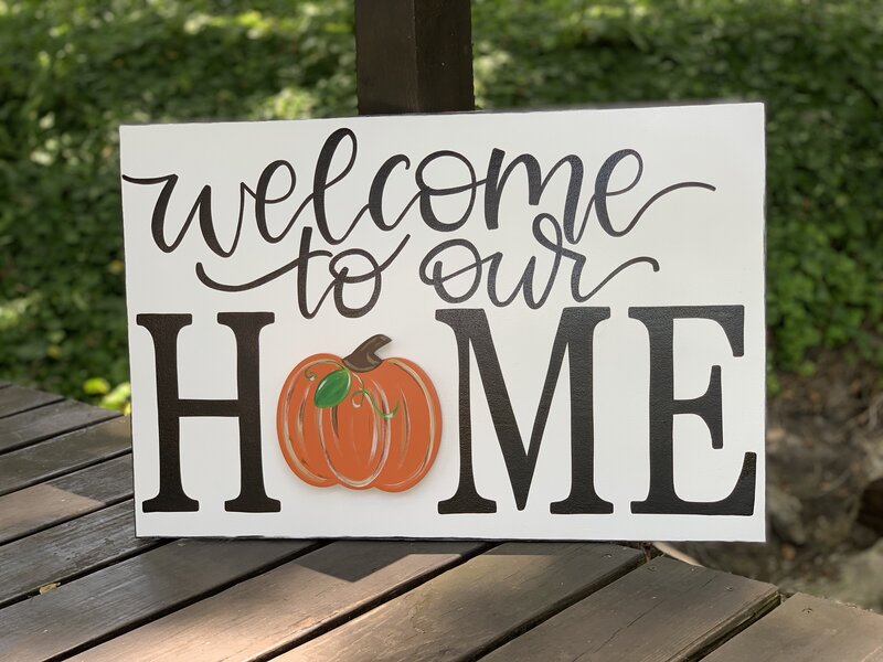Welcome to our home hand lettered in black on white rectangle wooden door hanger and orange pumpkin over the O in home