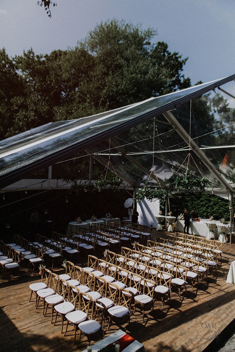 A breathtaking summer wedding tented celebration in Ramsey, NJ, featuring elegantly arranged long tables with wooden chairs under a clear canopy adorned with greenery and string lights.