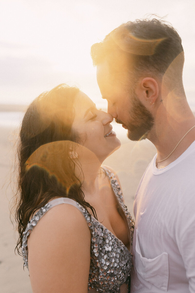 A couple leans in for a kiss on the beach at sunset, surrounded by warm golden light and a serene atmosphere.