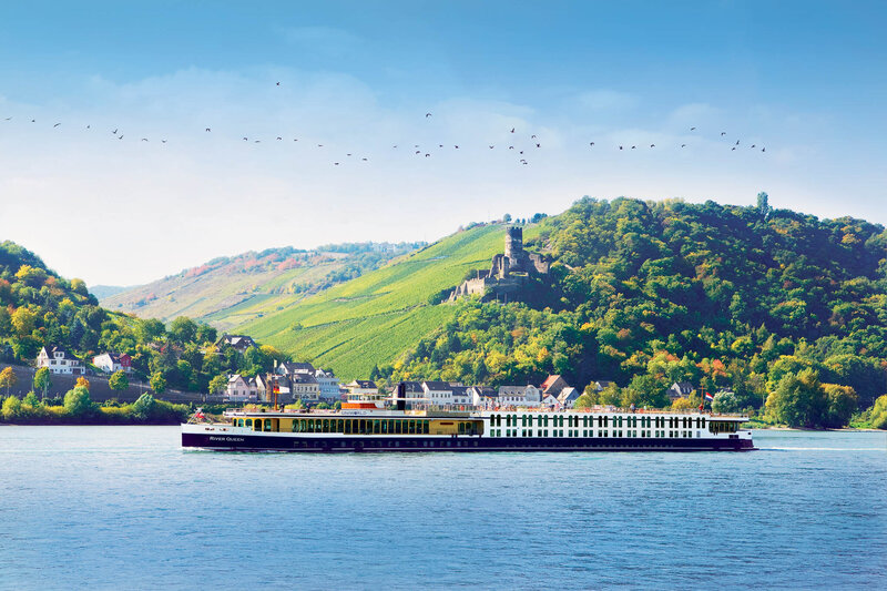 A long river cruise ship glides across a calm blue river, with green hills, vineyards, and a hilltop castle in the background under a clear sky with a flock of birds flying overhead.