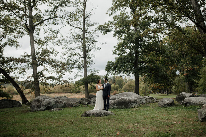 A couple stands surrounded by a sparse forest.