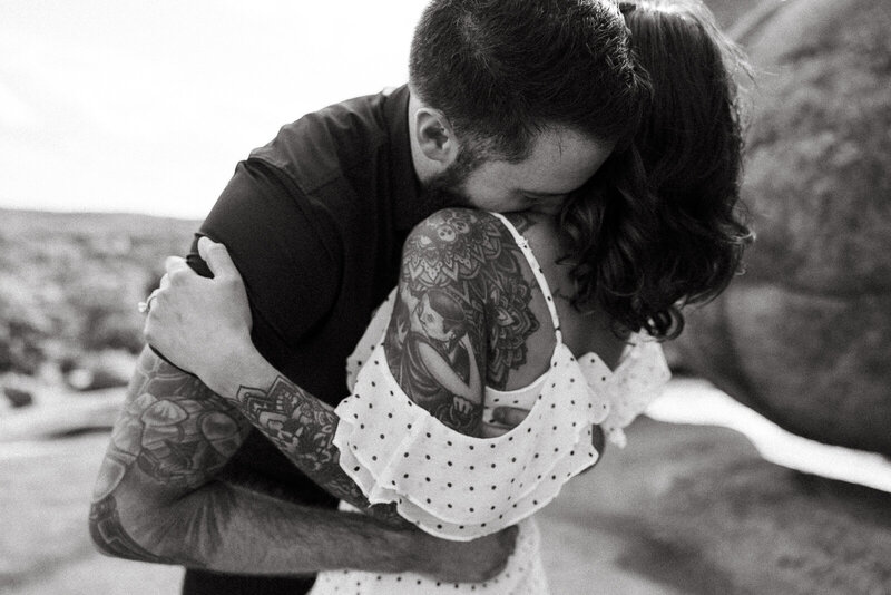 Couple with tattoos standing and cuddling during their engagement session with him kissing her neck. 