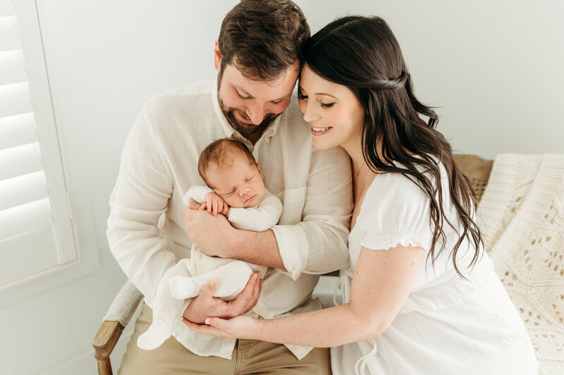 The newborn baby is in her fathers arms and both mum and dad is looking down and smiling during their newborn session in Atlanta.