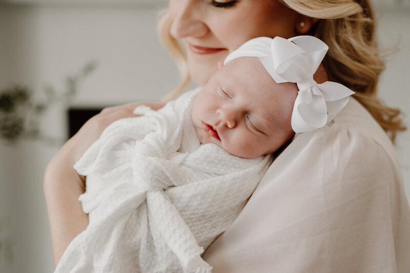 Motherhood photo of mom and newborn baby girl wearing white and mom looking down at baby girl in blanket resting on her shoulder.
