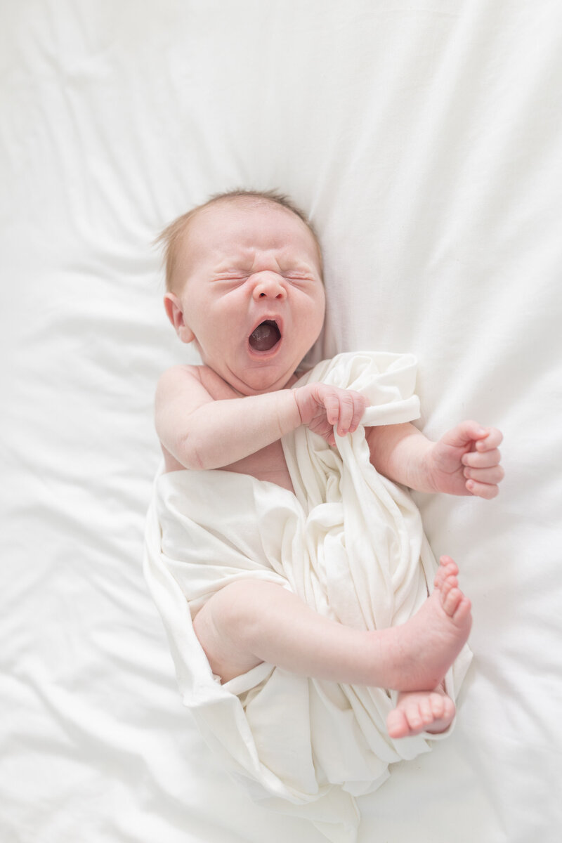 A newborn baby peacefully yawns while nestled in soft, white fabric. The gentle textures and bright, airy setting highlight the baby’s delicate features, capturing a pure and serene moment