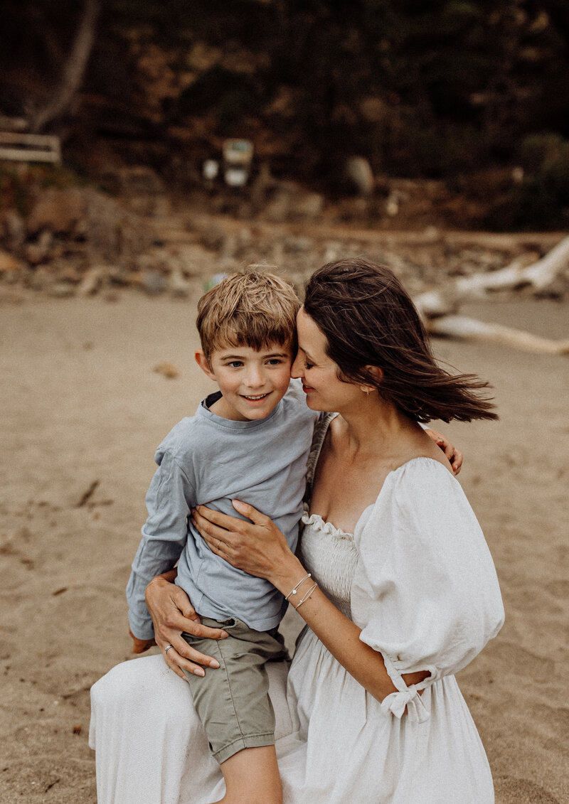 A mother wearing a white dress holding her son and posing for the camera as she is directed by Christchurch photographer Lucy Jane Photography