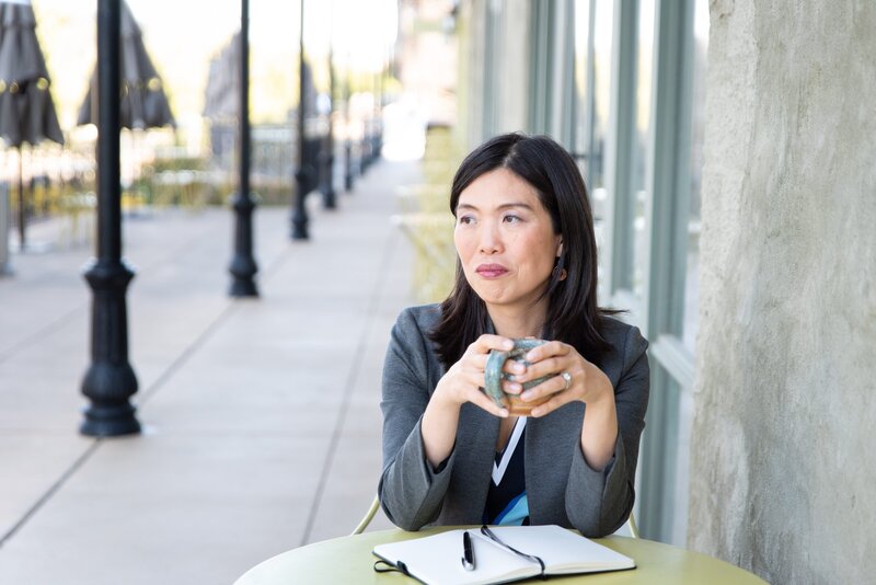 brand photo of woman sitting at a table outside a coffee shop, holding a coffee cup, smiling slightly and looking off to the left.