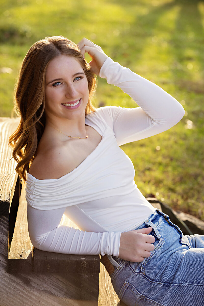 Smiling high school senior in jeans and white top seated on wooden steps of a rustic cabin at Anne Springs
