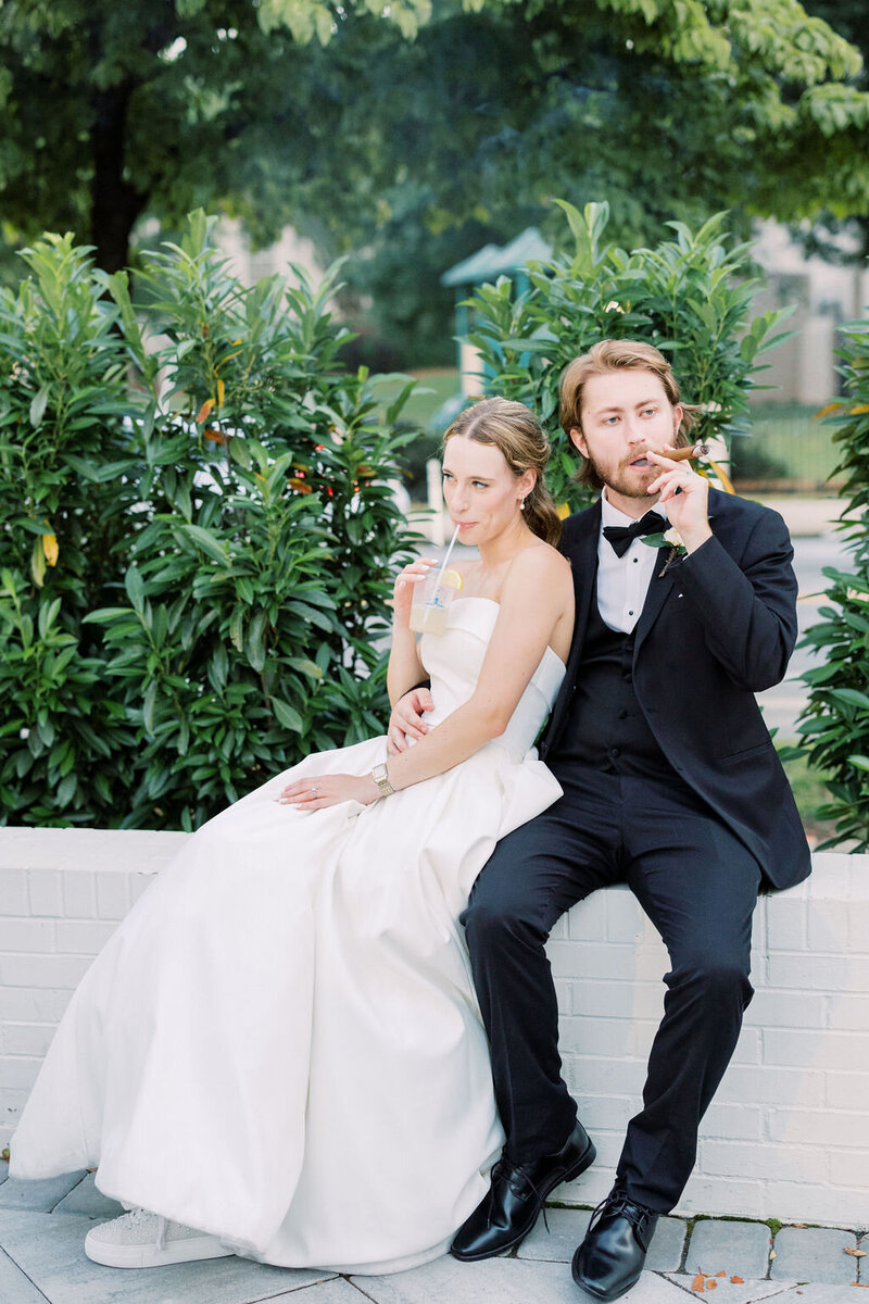Bride and groom sit on white wall with green plants in background