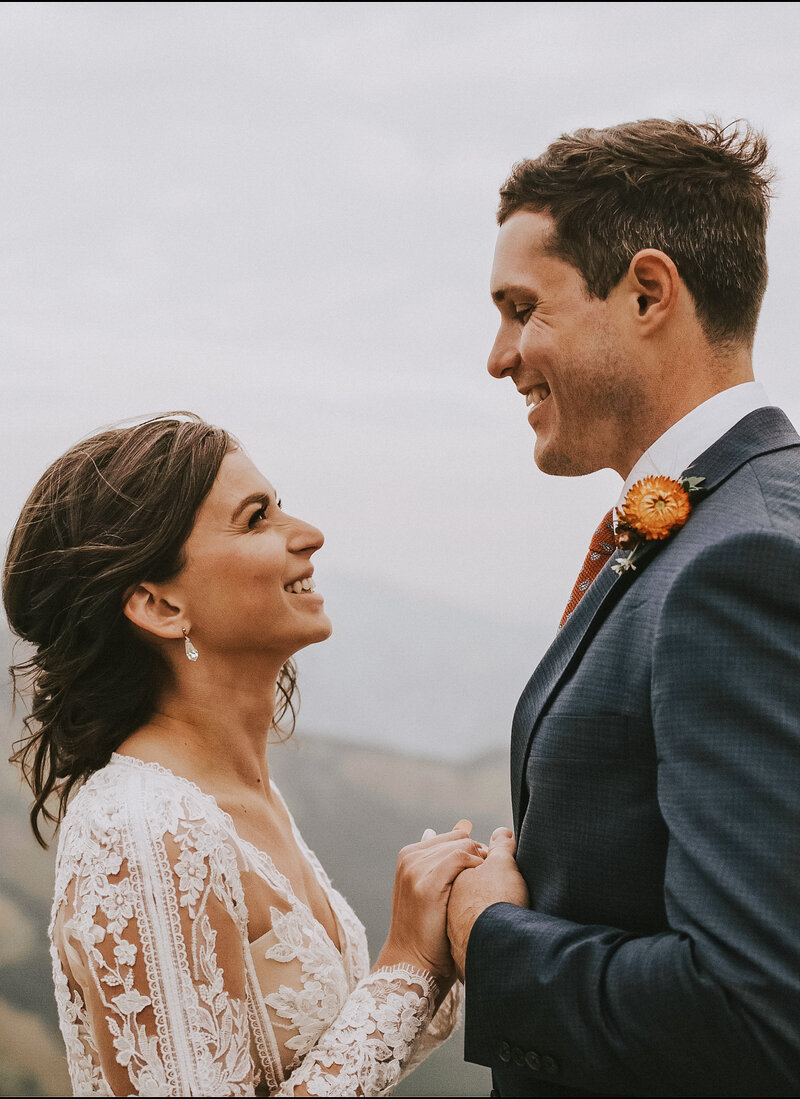 Bride and Groom shown standing smiling at one another on top of a mountain in Banff.