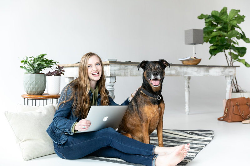 A woman sits with her laptop next to her dog on the floor in front of an antique desk in a brand photoshoot