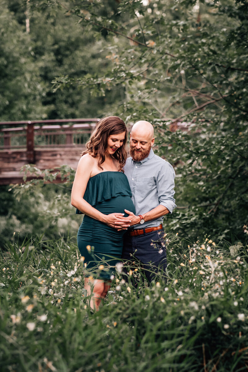 A maternity photo of a couple standing in a lush green setting. The woman is in a strapless green dress, and the man is holding her close as they both look down at her belly with smiles.