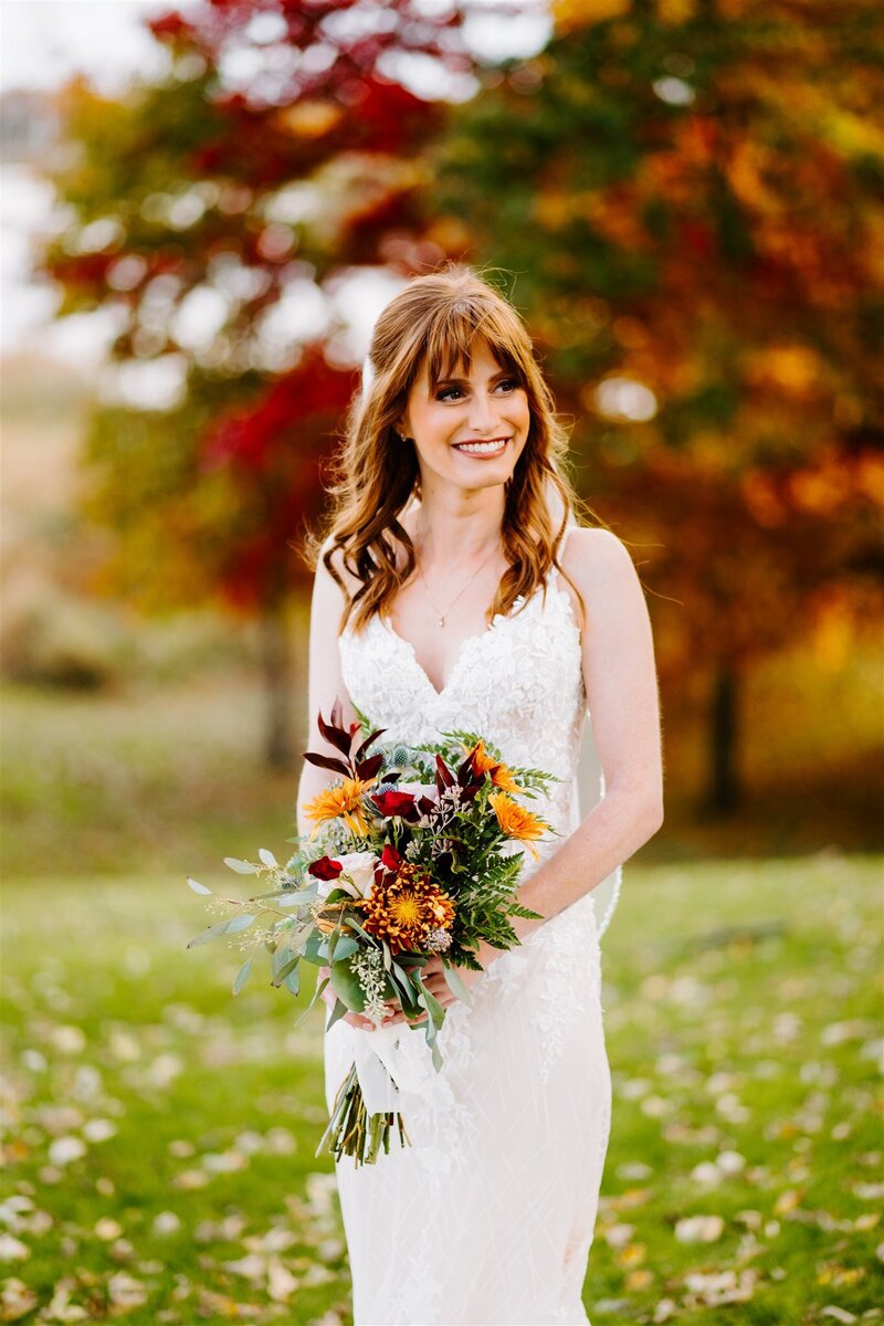 A joyful bride with long, wavy hair and a lace gown smiles warmly while holding a vibrant autumn bouquet with red, orange, and yellow flowers. She stands outdoors with a backdrop of colorful autumn leaves, creating a picturesque fall setting.