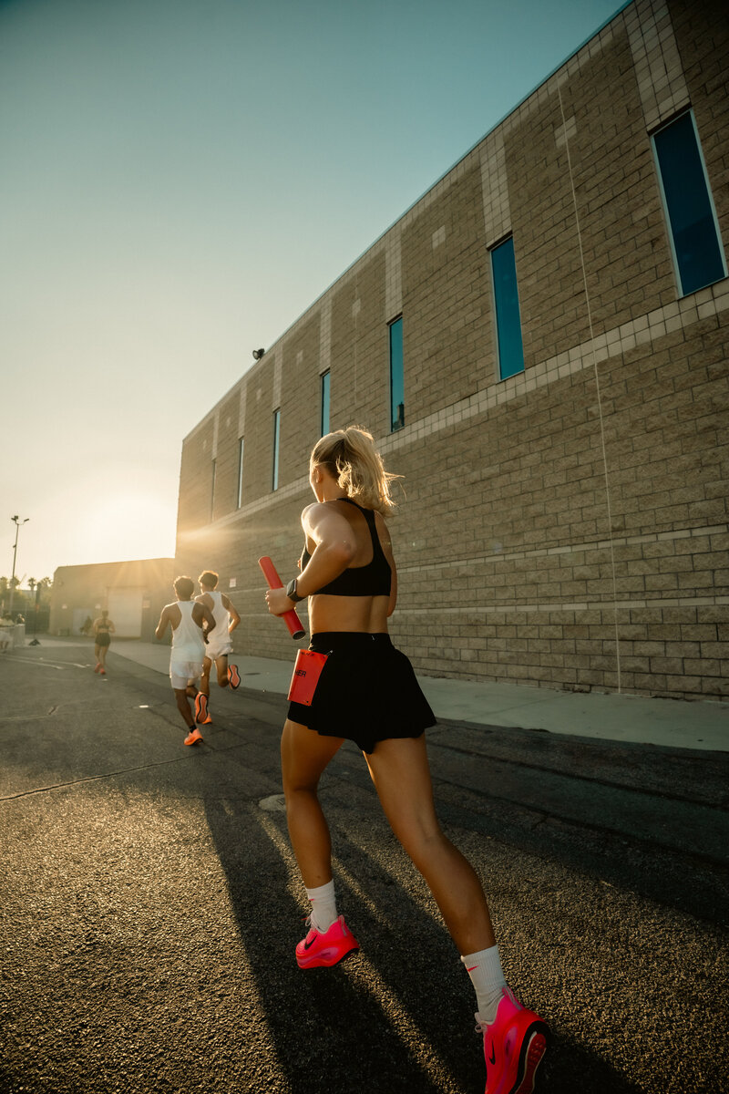 A HER Sports runner holding a baton while running with a group during a sunset training session beside a brick building.