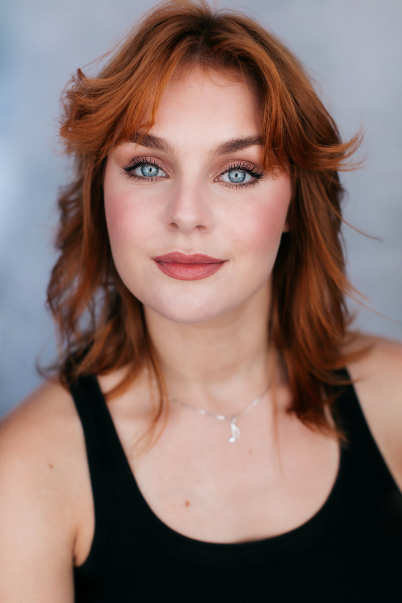 Casting headshot van performer met lang rood haar en zwarte top voor grijze achtergrond.