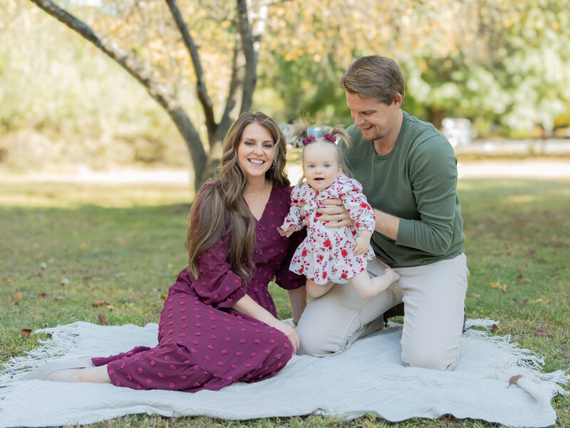 Family kneeling together for casual portrait on grass with autumn trees behind during an outdoor family session near Ramsey, NJ