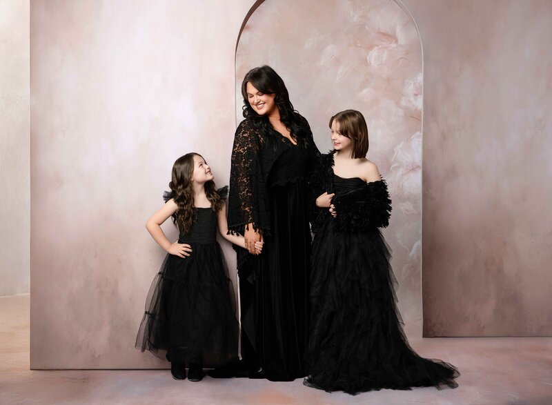 Mom and both daughters in black dresses posing for a portrait together. Mom is looking down and smiling at girls. Photographed by Atlanta photographer.