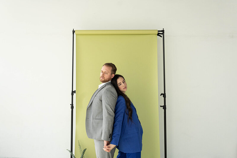 couple in pantsuits lean back to back in front of green backdrop