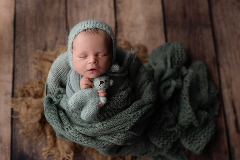 A cozy newborn portrait featuring soft sage knits and a beloved teddy companion—styled with rustic charm.