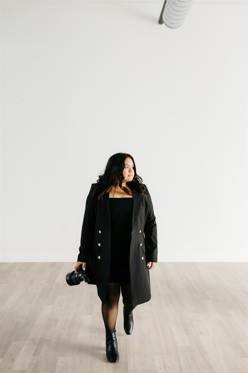 Virginia wedding photographer in all black dress, boots, and coat holding her camera and walking across a white studio space and looking to the side