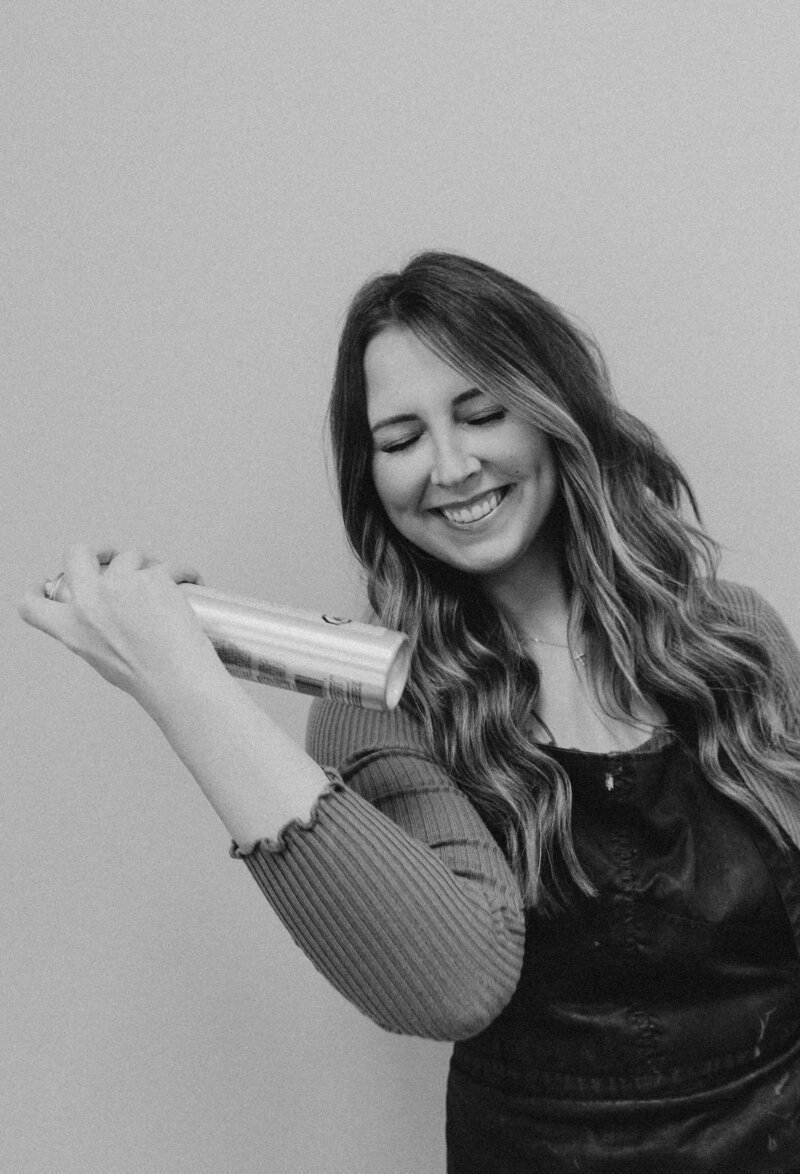 Black and white branding portrait of a smiling Midwest hairstylist spraying hairspray, captured during a professional branding session by a Michigan and Ohio branding photographer.