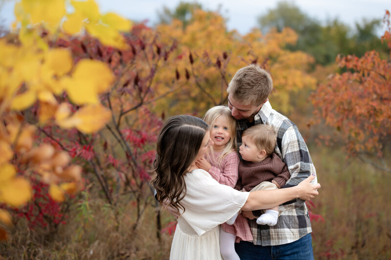 a smiling family hugs each other as they stand amidst yellow, red, and orange fall foliage in Minnesota