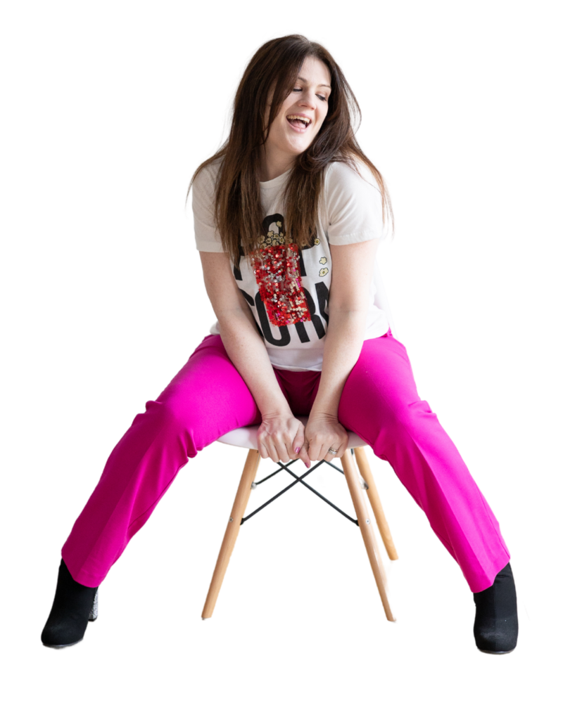 Emily wearing hot pink pants and a white t-shirt that says popcorn in black text with a sparkly image of popcorn on it. She is sitting on a white chair and has her eyes closed and her mouth open as is she was singing.
