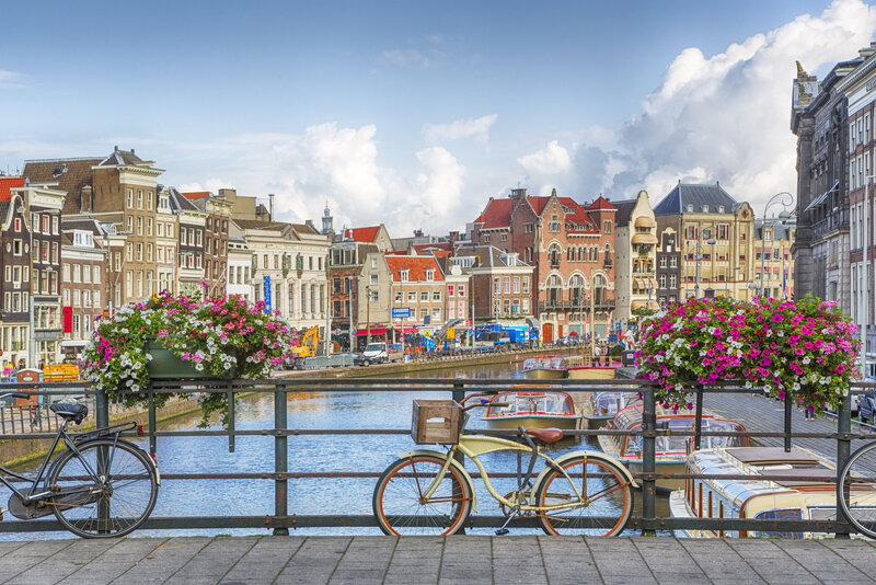 A serene view of the canals of Amsterdam from a bridge with a bike in front of it and flowers lining the bridge railings.