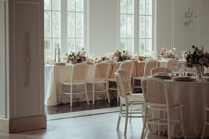 Reception tables by tall windows styled with neutral linens, florals, and white cane-back chairs.