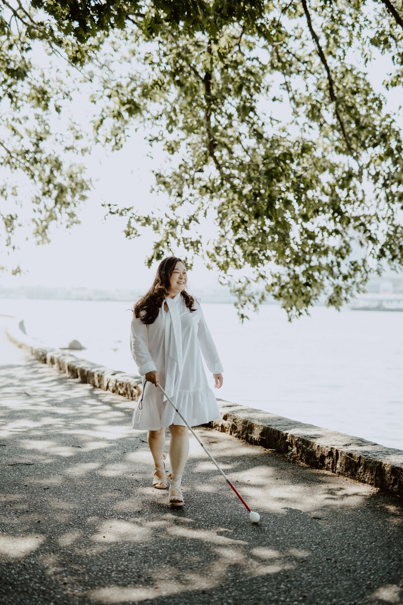 Anne, an Asian woman, walking outdoors along a shaded path beside a body of water. She is wearing a white long-sleeved dress that reaches just below her knees. She is holding a white cane in her right hand, which indicates that she is visually impaired. Her left hand is slightly raised, as if she is enjoying the breeze or the warmth of the sun. The background features a stone wall that borders the path and leafy tree branches overhead, casting dappled shadows on the ground. The scene exudes a sense of calm and natural beauty.