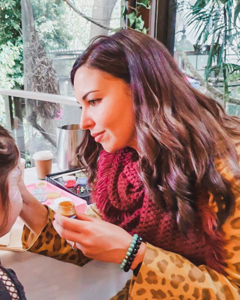 An image of Rania Fayad, CEO of Yellow Round Table face painting at a kids birthday party being held at the Melbourne Zoo. She is wearing a leopard print jacket, a burgundy scarf and an aqua and black beaded bracelet. Her hair is long, dark brown and wavy and she is smiling while holding a pot of gold paint and a paint brush.