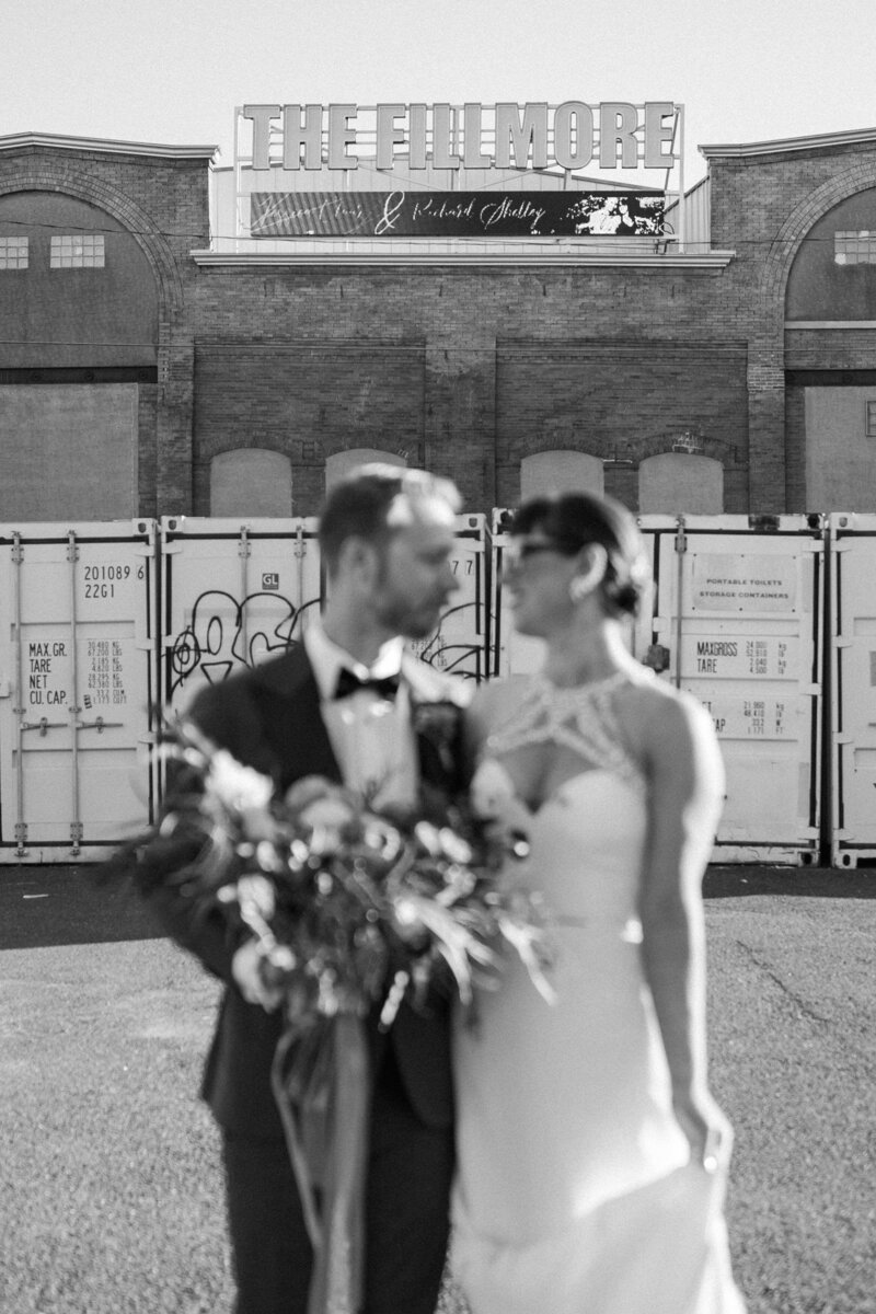 A couple dressed elegantly stands together with a bouquet, in front of a large sign for The Fillmore venue and storage containers.