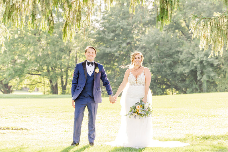 Newlyweds in a blue suit and lace dress hold hands while standing under a willow tree in a field for an Ann Arbor Wedding Photographer