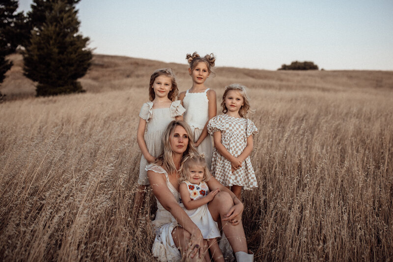 A mother and her four girls all wearing pretty neutral dresses sit in a golden El Dorado Hills field at sunset. 