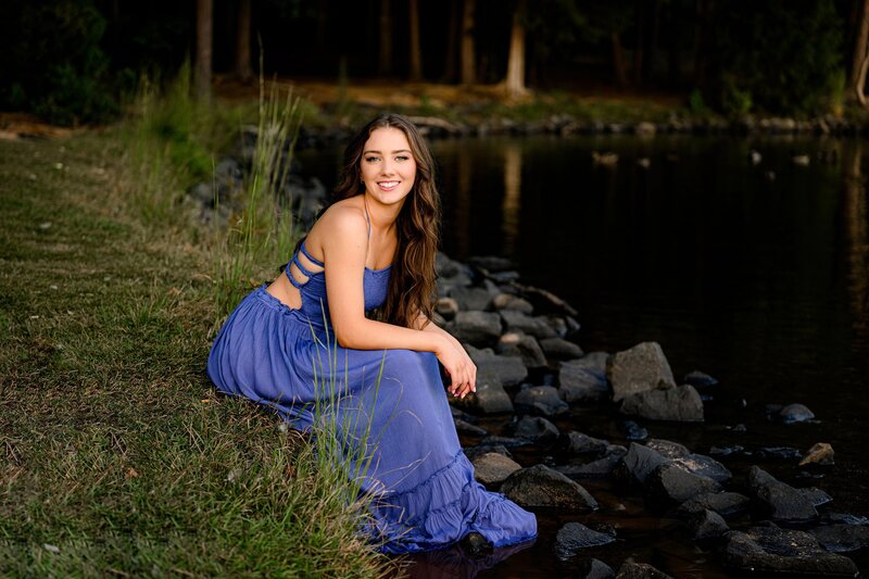 senior sits on the edge of a lake with her elbows on her knees during her grad session wearing a blue dress