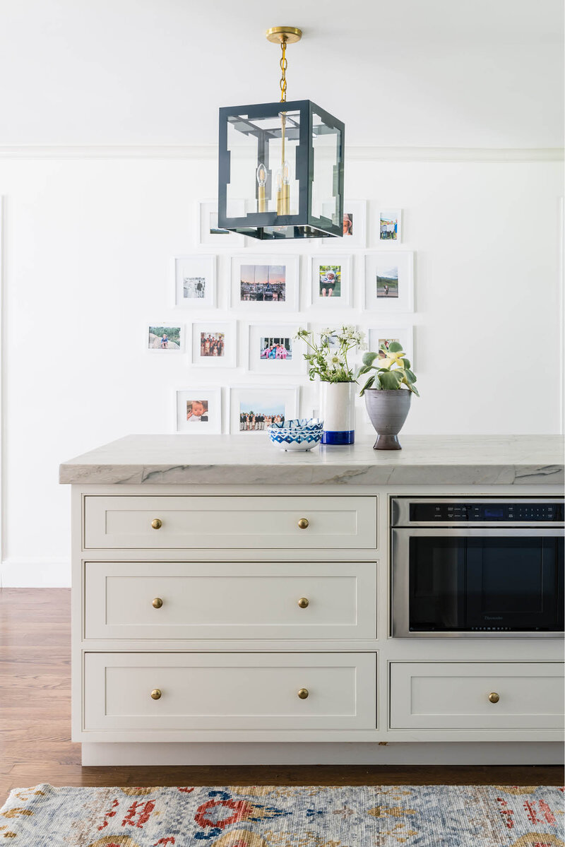 Explore this simple yet elegant white kitchen featuring a stacked marble island beneath a custom blue lantern. A collection of family photos adds a personal touch to the white walls, while a colorful area rug brings warmth to the space.