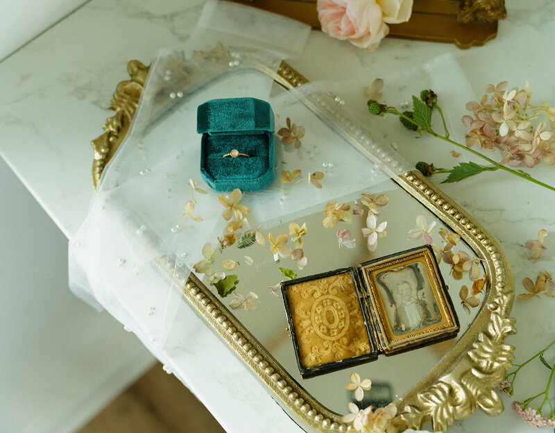 Wedding day shoot, details shot. A wedding ring in a dark green velvet ring box is sitting on top of a vintage gold mirror with little flowers around it.
