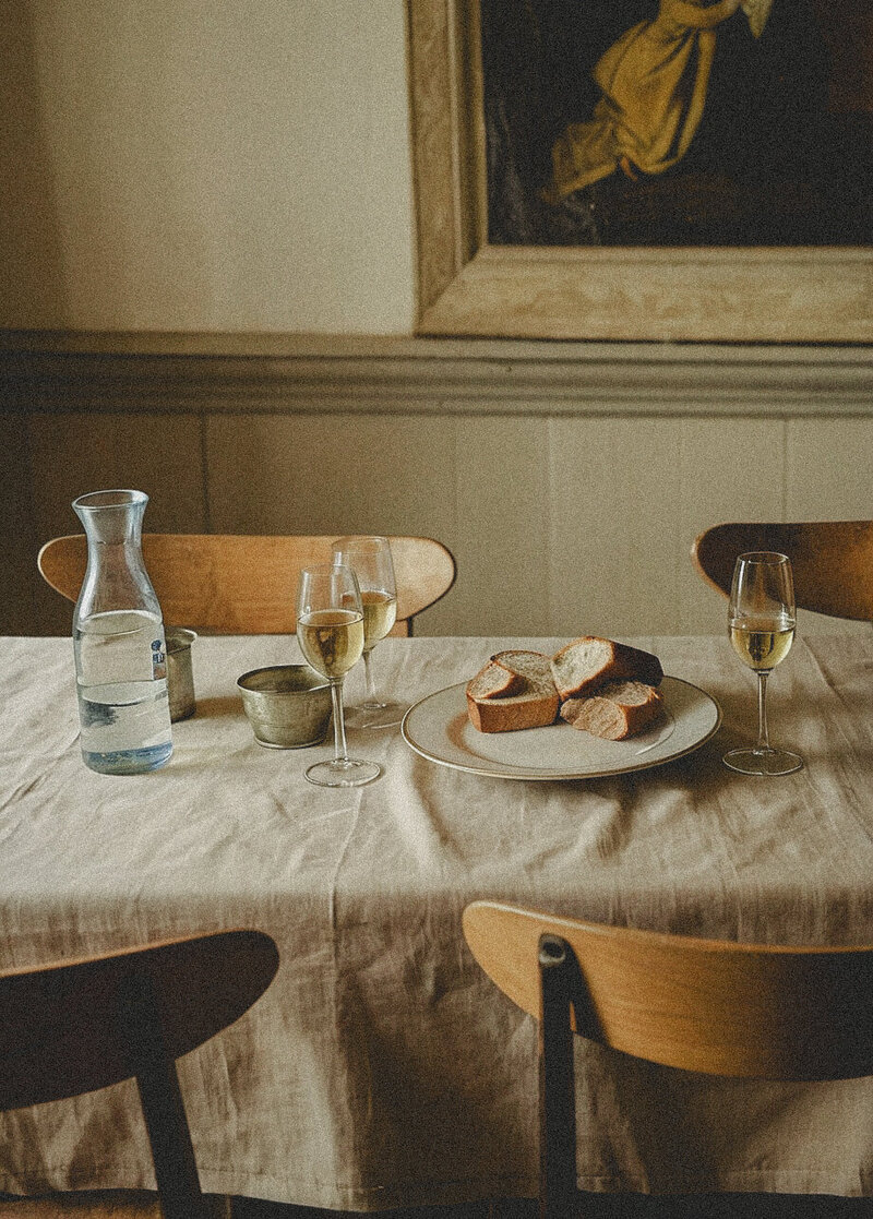 Farmhouse kitchen table with linen flax colored tablecloth with plate of bread and glasses of wine and a carafe of water and tins on top of the table with antique portrait on wall behind table