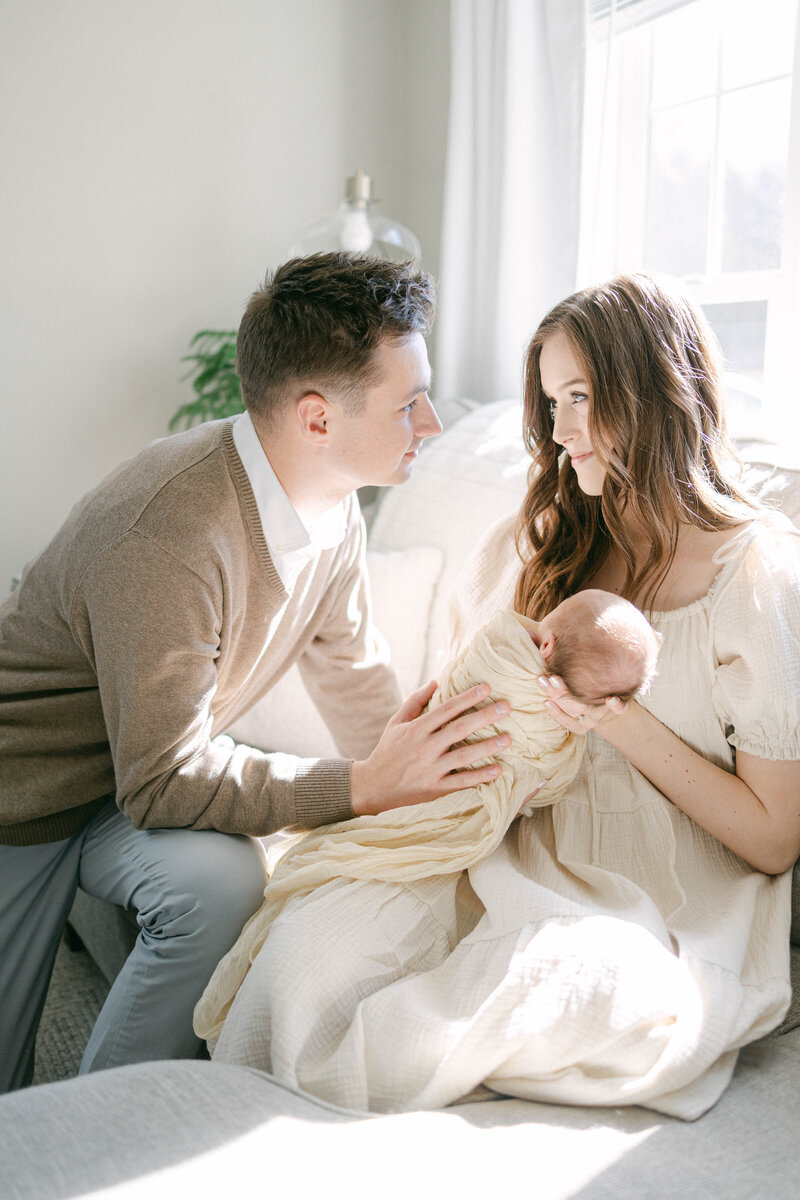 man and women on the couch looking at each other while holding newborn baby with  bright daylight pouring in through windows knoxville tennessee newborn photographer