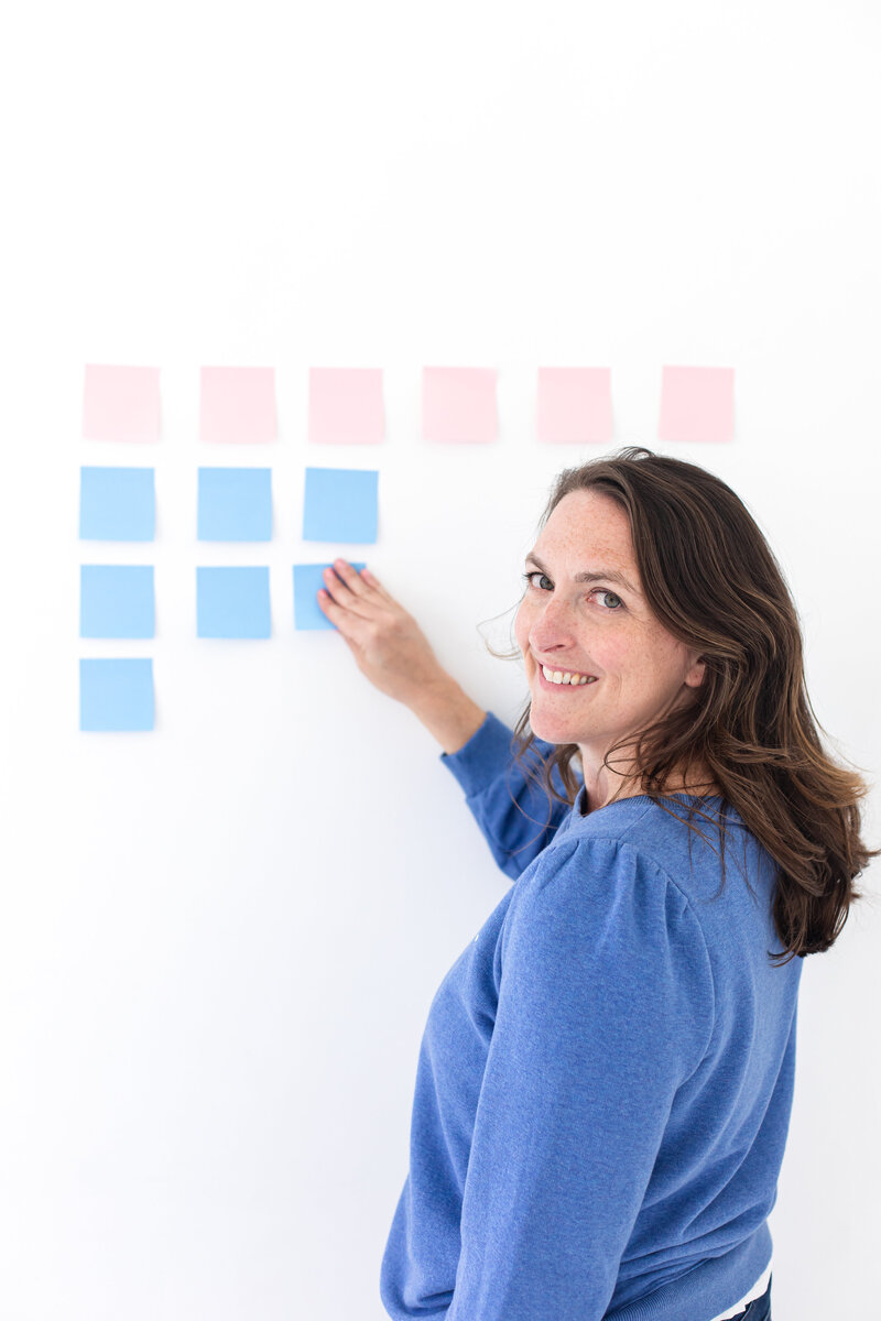 Social media strategist and course creator katy miller, dressed in a blue jacket, holds light-colored post it papers in her right hand to stick them on the white wall