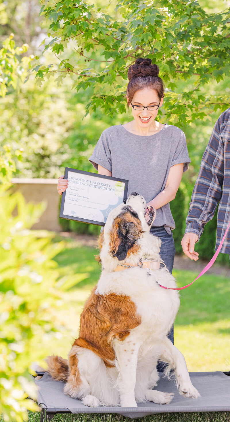 Woman smiling at her St. Bernard, holding up a graduation certificate for completing training, outdoors in Canton, MI