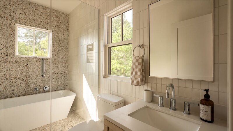 Sunlit bathroom with white and tan tones, soaking tub inside glass-enclosed tiled shower, small vanity with square mirror, checkered towel, and window over toilet.
