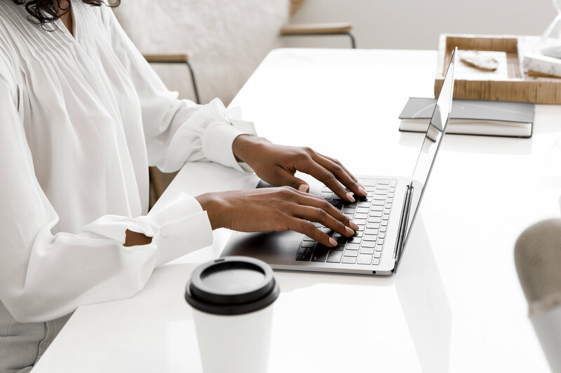Close-up of woman’s hands typing on a laptop with coffee on a white desk
